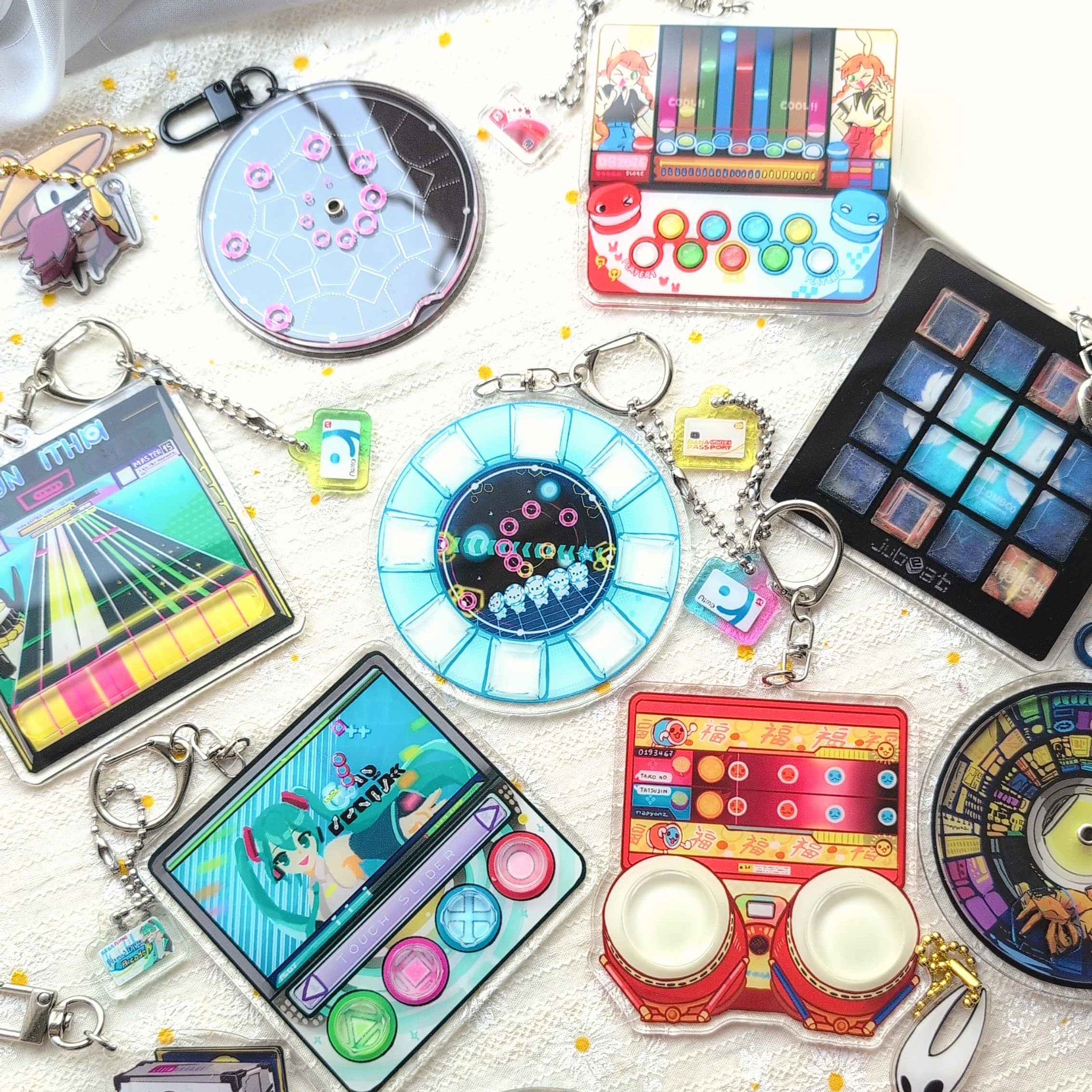 A collection of colorful mini keychains and charms featuring retro music instruments, game screens, and cartoon designs, arranged on a white fabric background.