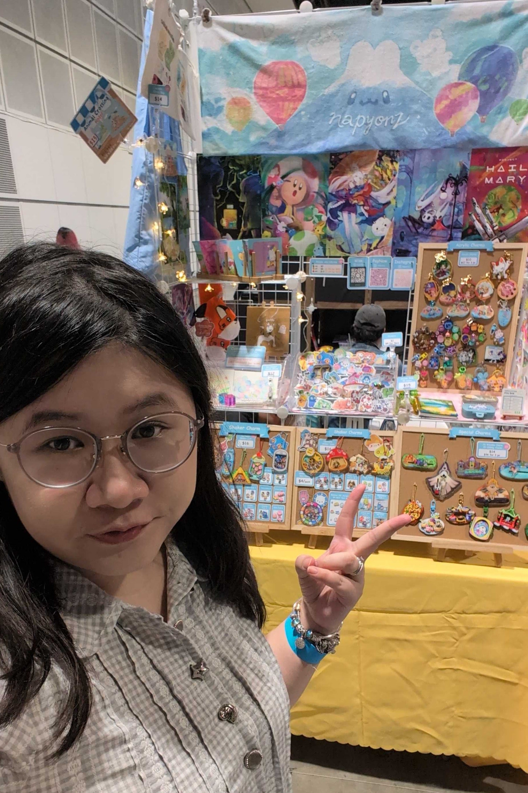 A woman with glasses and dark hair poses in front of a colorful anime merchandise booth, making a peace sign. The booth features a blue banner with hot air balloon illustrations, shelves of small stickers, pins, and other collectibles, and a yellow tablecloth underneath.